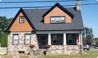 A two-story house with a stone facade, brown siding, and a black roof crafted by a skilled roofer in Riverdale NJ. The front porch features white columns and two potted plants with red flowers on each side of the steps. A lush grassy lawn surrounds the house, and a car is parked nearby.