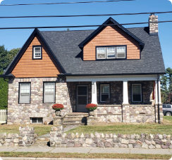 A stone house with a dark roof and orange wood accents, featuring a small porch. Two red potted plants adorn the entrance, and a stone wall borders the front yard. Power lines run across the top of the image, maintained by a skilled roofer in Riverdale NJ, with trees in the background.