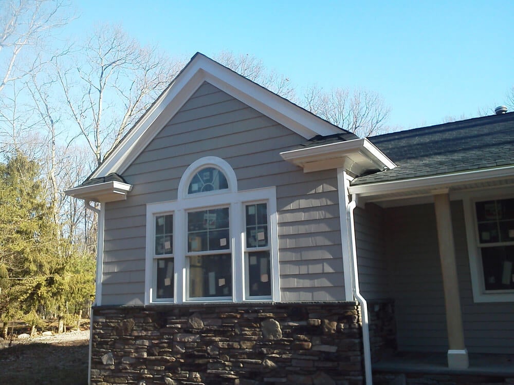A house exterior in Riverdale, NJ, displays a single-story section with gray siding and a stone base. The front features a white-trimmed window with an arched design above it. Bare trees and a clear blue sky can be seen in the background, highlighting the expertise of local roofing services.
