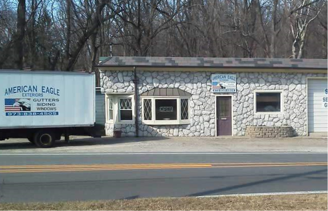 A building with a stone exterior houses "American Eagle Exteriors," a prominent roofing contractor in Riverdale, NJ. A sign with their logo adorns the entrance, and a branded truck is parked on the left. A road runs in front, lined with trees in the background.