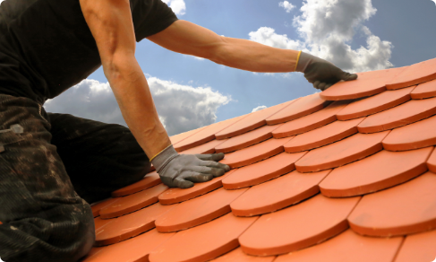 A roofing contractor in Riverdale NJ is diligently installing reddish-brown shingles on a roof under a partly cloudy sky. The shingles have a rounded, scalloped shape and fit together to form a neat pattern, demonstrating the contractor's skill and precision.
