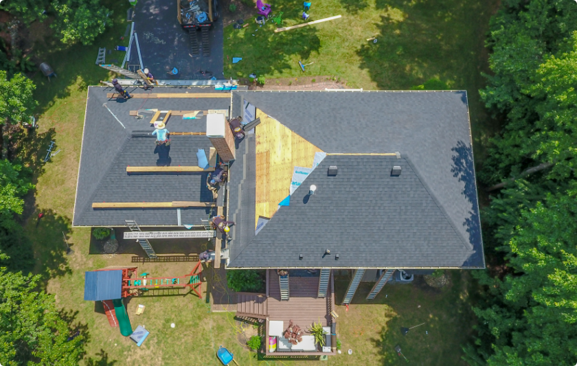 Aerial view of a house roof undergoing repair by a skilled roofer in Riverdale, NJ. Several workers are installing new shingles and wooden panels. Ladders line the sides, with construction materials scattered around the property. Surrounding trees provide a lush backdrop.