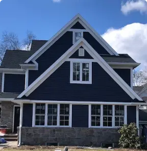 A two-story house with dark blue siding in Riverdale, NJ, and white trim stands under a partly cloudy sky. The front features multiple windows and a gabled roof, crafted by a renowned roof company in Riverdale, NJ. A small shrub is planted near the stone foundation.