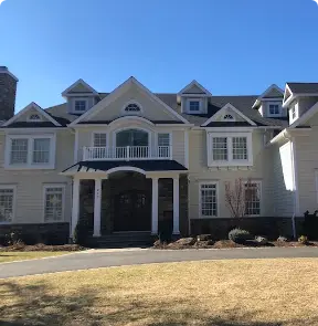 A large, two-story white house with expertly installed siding in Riverdale NJ boasts multiple gables and a dark front door. A small balcony graces the entrance, all surrounded by a well-maintained lawn under the clear blue sky.