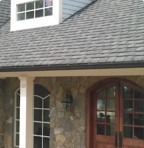 A house with a stone facade, featuring a wooden double door with glass panels and arched design. A white column supports a small overhang, while a large window sits to the left. The shingled roof, expertly crafted by a roofing contractor in Riverdale NJ, boasts an elegant dormer window above.