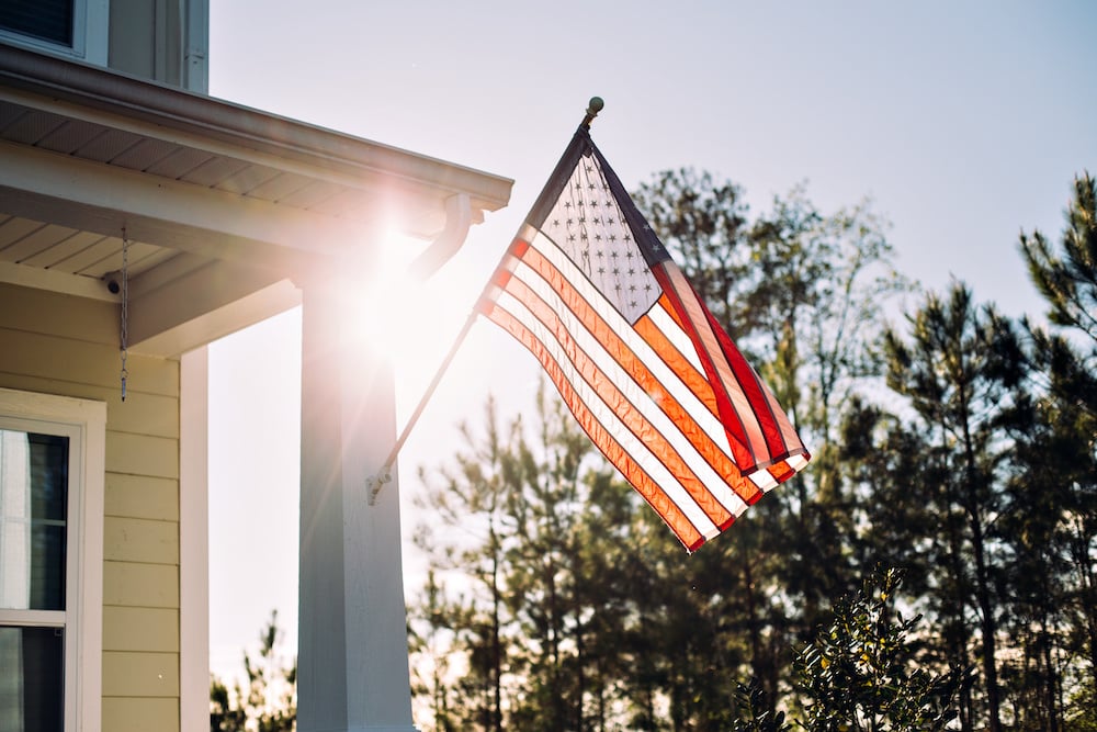The image shows an American flag waving in the sunlight, attached to a house. In the background, a clear blue sky and tall trees grace the scene. The sun creates a flare effect near the edge of the roof, hinting at expert roofing craftsmanship by a trusted contractor in Riverdale NJ.