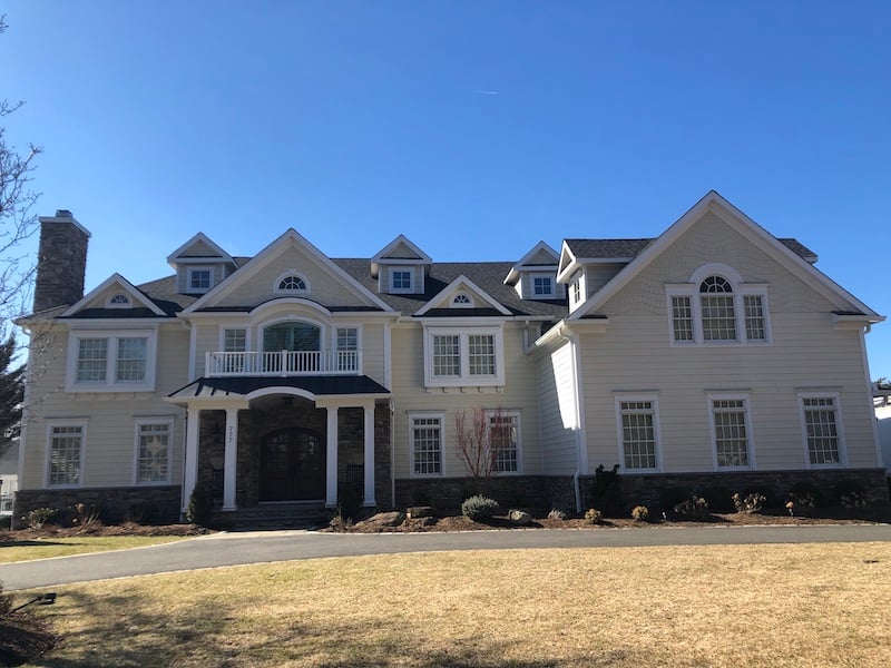 A large two-story house with white siding and a stone facade, expertly crafted by a roofing contractor in Riverdale NJ. It features multiple dormer windows, a balcony above the entrance, and a circular driveway. The sky is clear and blue, accentuating the well-maintained front lawn.