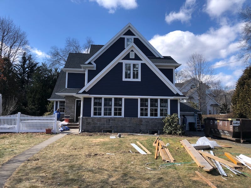 A two-story house with a blue facade and white trim sits under a partly cloudy sky in Riverdale, NJ. Construction materials from the local roof company are scattered on the lawn. A white picket fence and trees complement this charming scene.