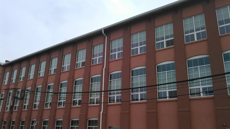 A large three-story brick building with multiple windows in each row. The facade features reddish-brown walls with a white drainage pipe running vertically, a testament to the expert gutter services in Riverdale NJ. Overhead power lines are visible in the foreground under a cloudy sky.