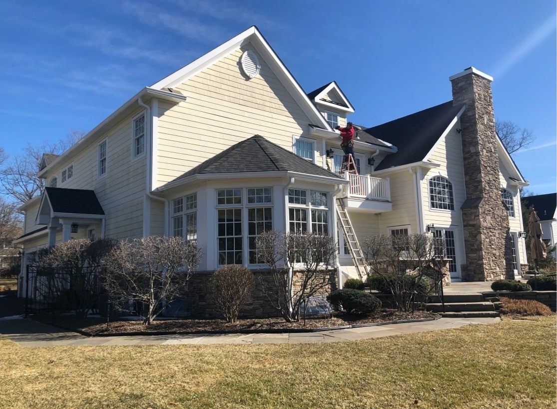 A person stands on a ladder cleaning or working on the second-story windows of a large beige house with white trim, stone accents, and a well-maintained yard under a clear blue sky.