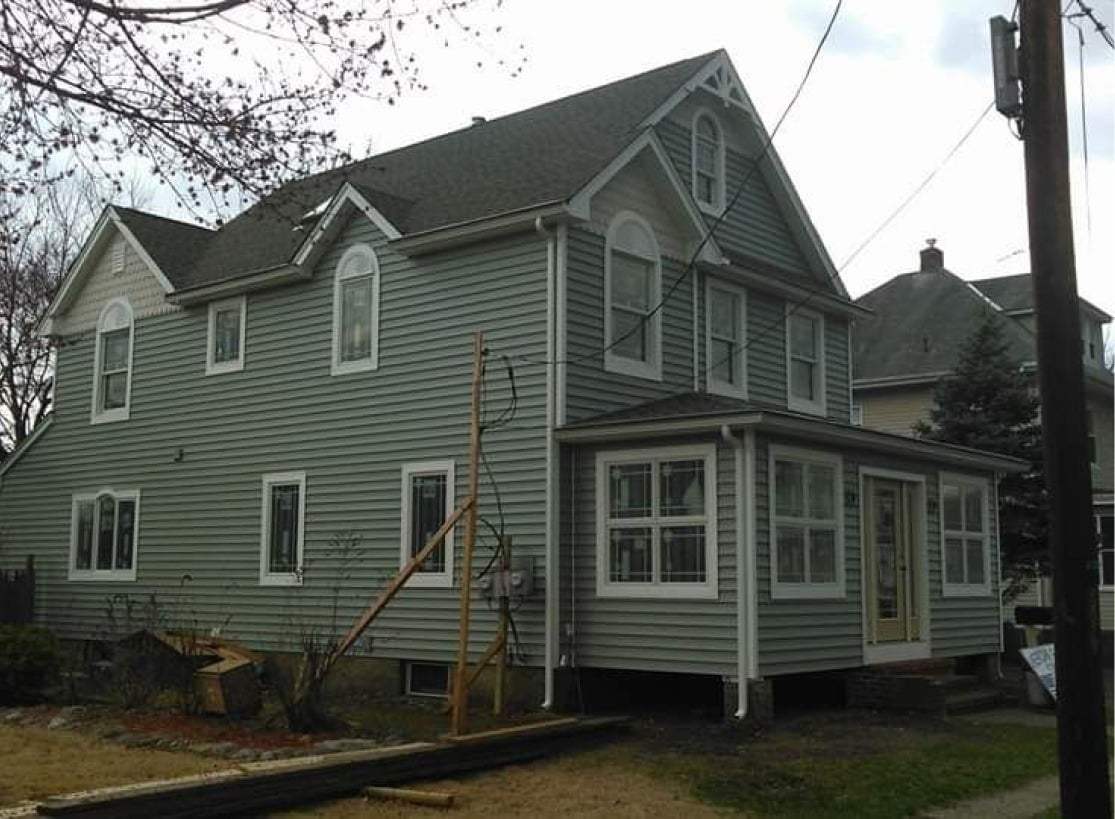 A two-story house with gray siding, white trim, multiple windows, and a small front porch. The yard is bare with minimal landscaping, and there is a utility pole and some trees nearby.
