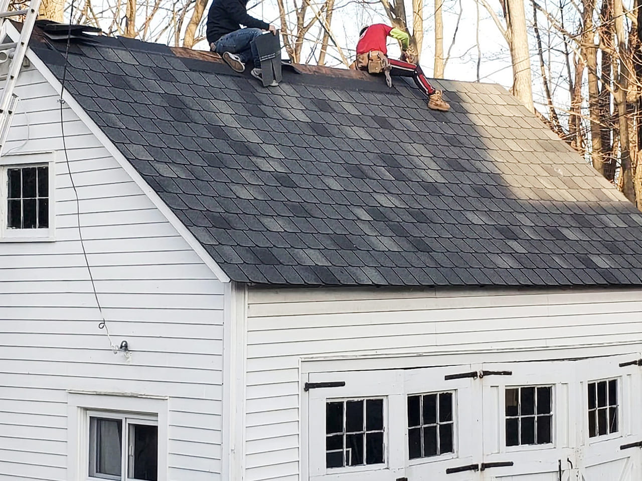 Two people are working on the sloped shingle roof of a white building with garage doors. One person is kneeling, and the other is sitting, both near the roof peak. Trees without leaves are visible in the background.