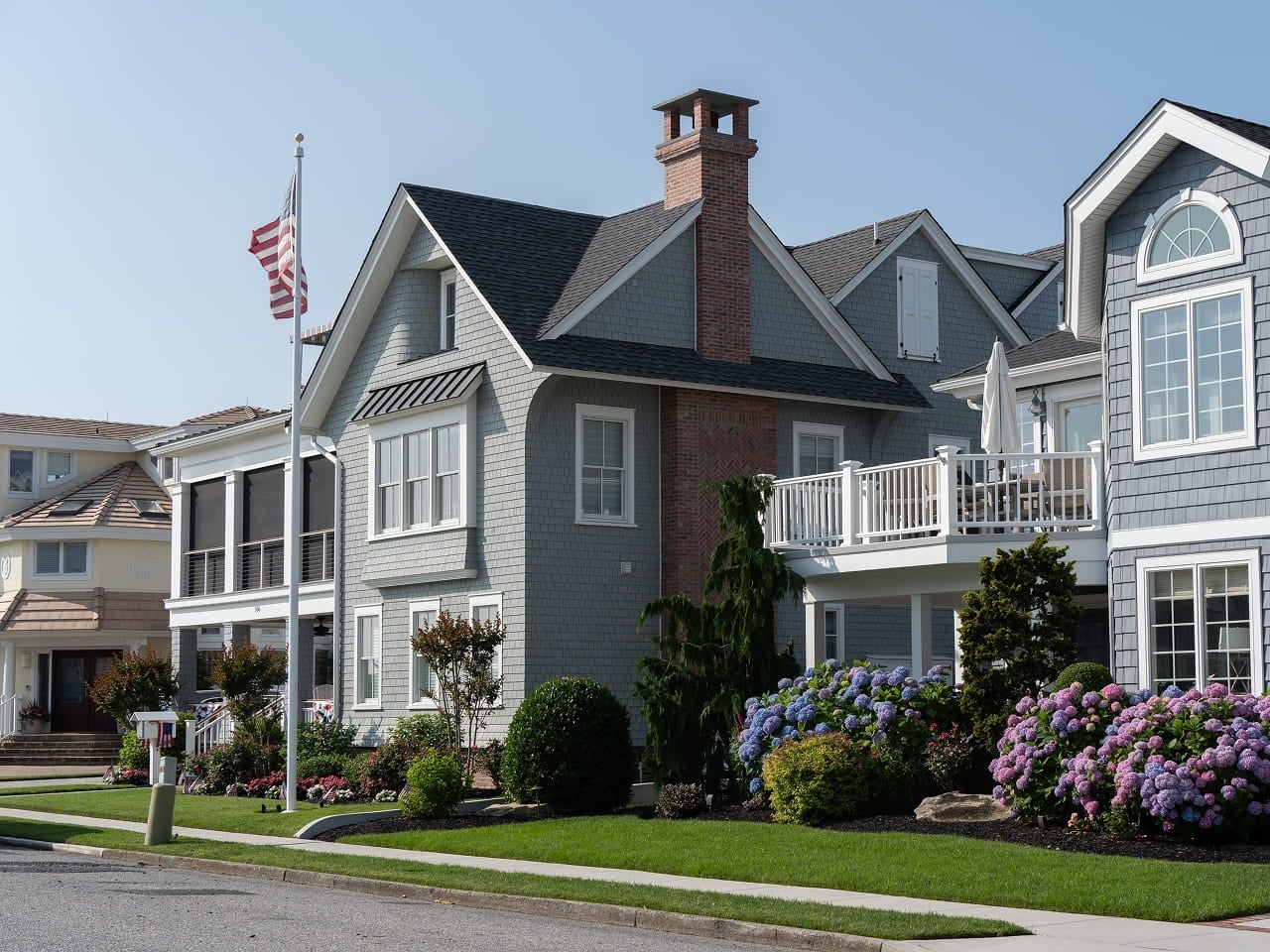 A large, gray two-story house with white trim, a brick chimney, a balcony, and a well-kept garden with blooming flowers, sits on a sunny suburban street. An American flag flies from a flagpole in the front yard.