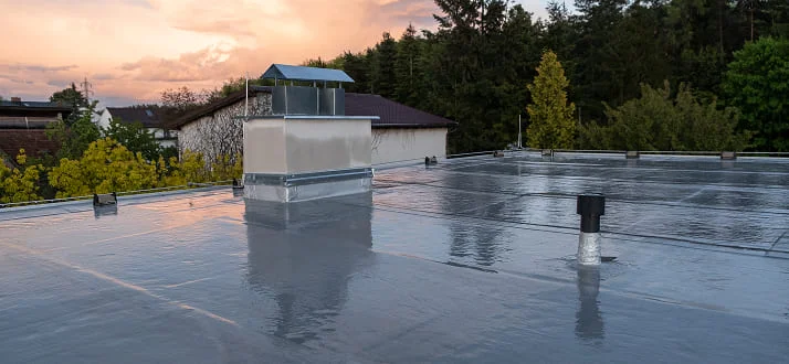 A flat roof covered with a shiny, waterproof membrane, featuring vents and a chimney, with trees and buildings in the background at sunset.