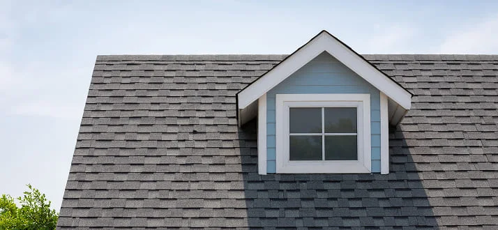 A close-up of a gray shingled house roof with a small dormer window, blue siding, and white trim, set against a clear sky.