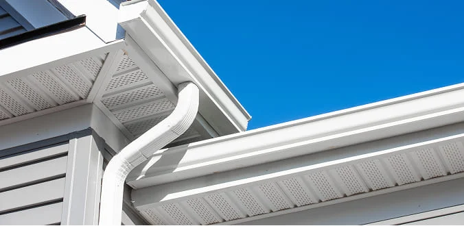 White rain gutter attached to the edge of a house roof against a bright blue sky, showing clean soffit and siding details.