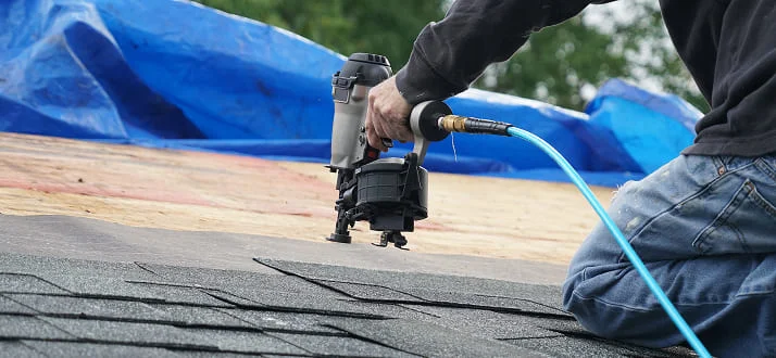 A person uses a pneumatic nail gun to install asphalt shingles on a roof. Blue tarps and exposed wooden sheathing are visible in the background.