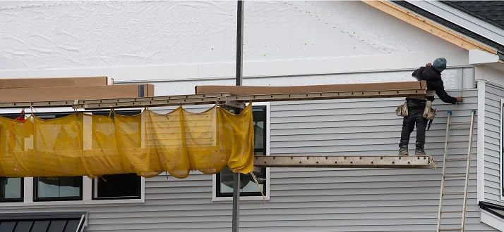 A construction worker stands on scaffolding, installing siding on a house. The worker is near the roof and wears a tool belt. A yellow safety net is attached to the scaffolding.