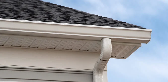 A close-up view of a house roof corner with dark shingles, white soffit, fascia, and a white rain gutter with a downspout against a blue sky.